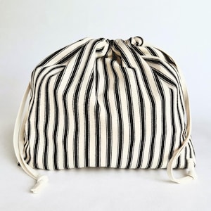 Pode incluir: Uma bolsa com cordão com um padrão de listras pretas e brancas. A bolsa é feita de tecido e possui um fecho de cordão de cor creme. As listras são verticais e uniformemente espaçadas.