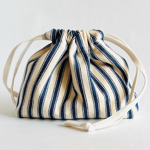 May include: A small drawstring bag featuring a cream-coloured background with navy blue vertical stripes. It has a white drawstring closure at the top. The bag is made of fabric and is suitable for storing or carrying small items.