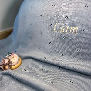 Peut inclure: Une couverture tricotée bleu clair avec un motif à œillets répétitif et le prénom Liam brodé en fil jaune clair. Un pelote à épingles ronde avec des épingles colorées et une paire de ciseaux en métal sont posées sur le tissu.