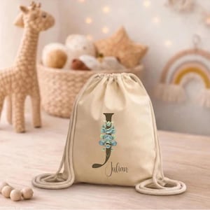 Könnte beinhalten: Ein beigefarbener Kordelzugrucksack mit einem großen dunkelbraunen Buchstaben J, der mit einer Illustration eines Eukalyptuszweigs im Aquarellstil verziert ist. Der Name Julian ist in dunkelbrauner Schreibschrift unter dem Buchstaben gedruckt.