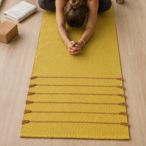 Puede incluir: Una esterilla de yoga rectangular de color amarillo mostaza con una textura tejida y filas horizontales de flecos marrones. La esterilla está colocada sobre un suelo de madera clara, proporcionando una superficie plana para ejercicios.