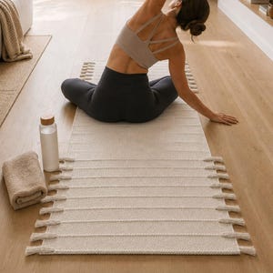 May include: A rectangular beige yoga mat featuring a textured surface with horizontal ridges and decorative fringe tassels along both long sides. The mat is placed on a wooden floor alongside a rolled beige towel and a clear water bottle with a wooden lid.
