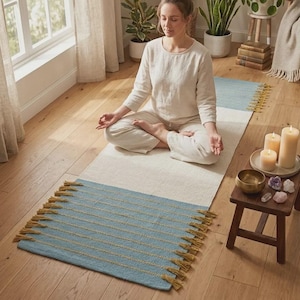 May include: A rectangular yoga mat featuring a central cream-colored section flanked by two light blue ends. The blue sections are decorated with horizontal rows of thin, mustard-yellow thread detailing and finished with matching mustard-yellow fringe.