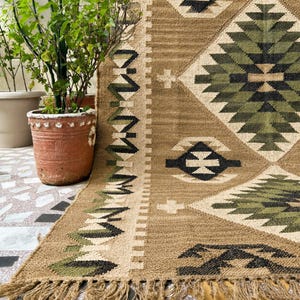 May include: A woven area rug featuring a tan base color with a large central diamond pattern in shades of olive green, black, and cream. The rug includes geometric border motifs, a fringed edge, and a textured surface with a visible weave pattern.