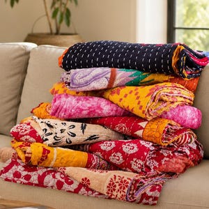May include: A stack of folded kantha quilts featuring various patterns and colours, including dark blue, yellow, pink, red, and white. The quilts display intricate stitching details and diverse floral and geometric prints layered on top of each other.
