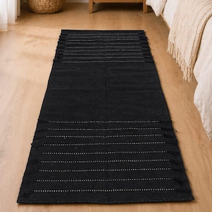 May include: A rectangular black runner rug featuring a series of thin, horizontal white dashed lines spaced evenly across its surface. The rug has short, knotted fringe details along both of its shorter ends, creating a textured finish.