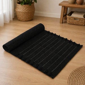 May include: A rectangular black rug featuring a subtle speckled texture and thin, evenly spaced horizontal lines. The rug is partially rolled at one end and finished with short, decorative fringe tassels along the side edges of the fabric.