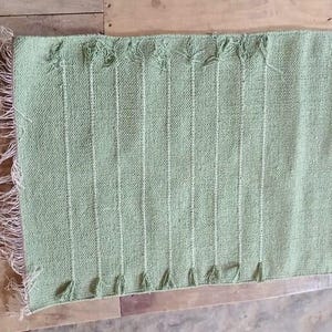 May include: A rectangular sage green woven runner featuring a textured surface with vertical raised lines and a fringed edge on one side. The fabric has a consistent light green hue and a visible weave pattern throughout the entire length of the piece.
