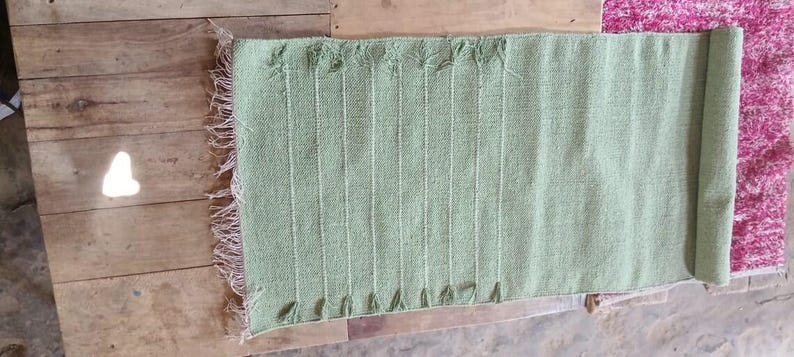 May include: A rectangular sage green woven runner featuring a textured surface with vertical raised lines and a fringed edge on one side. The fabric has a consistent light green hue and a visible weave pattern throughout the entire length of the piece.