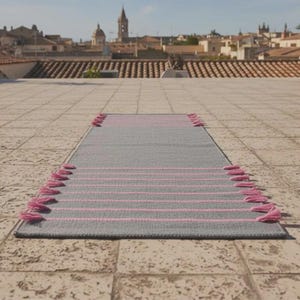 May include: A rectangular runner rug featuring a solid gray base with horizontal pink stripes and pink tassels attached along the two shorter sides. The rug is positioned flat on a tiled surface with a town skyline visible in the background.