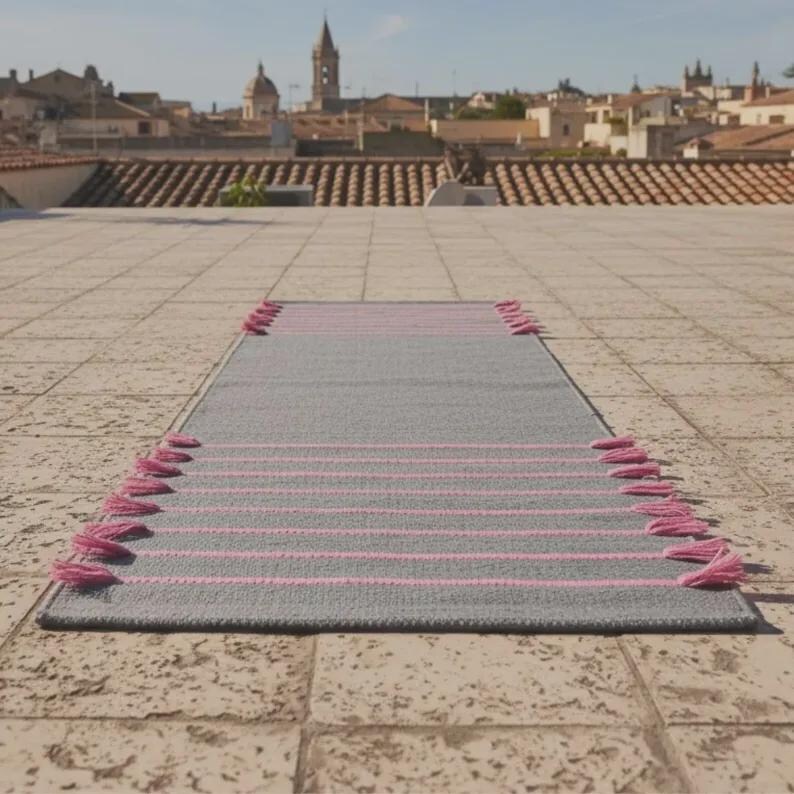 May include: A rectangular runner rug featuring a solid gray base with horizontal pink stripes and pink tassels attached along the two shorter sides. The rug is positioned flat on a tiled surface with a town skyline visible in the background.