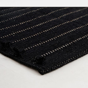 May include: A rectangular black woven rug featuring a series of parallel horizontal lines in a light beige or off-white color. The texture of the rug appears thick and coarse, with small knotted fringe details visible along the edge of the fabric.