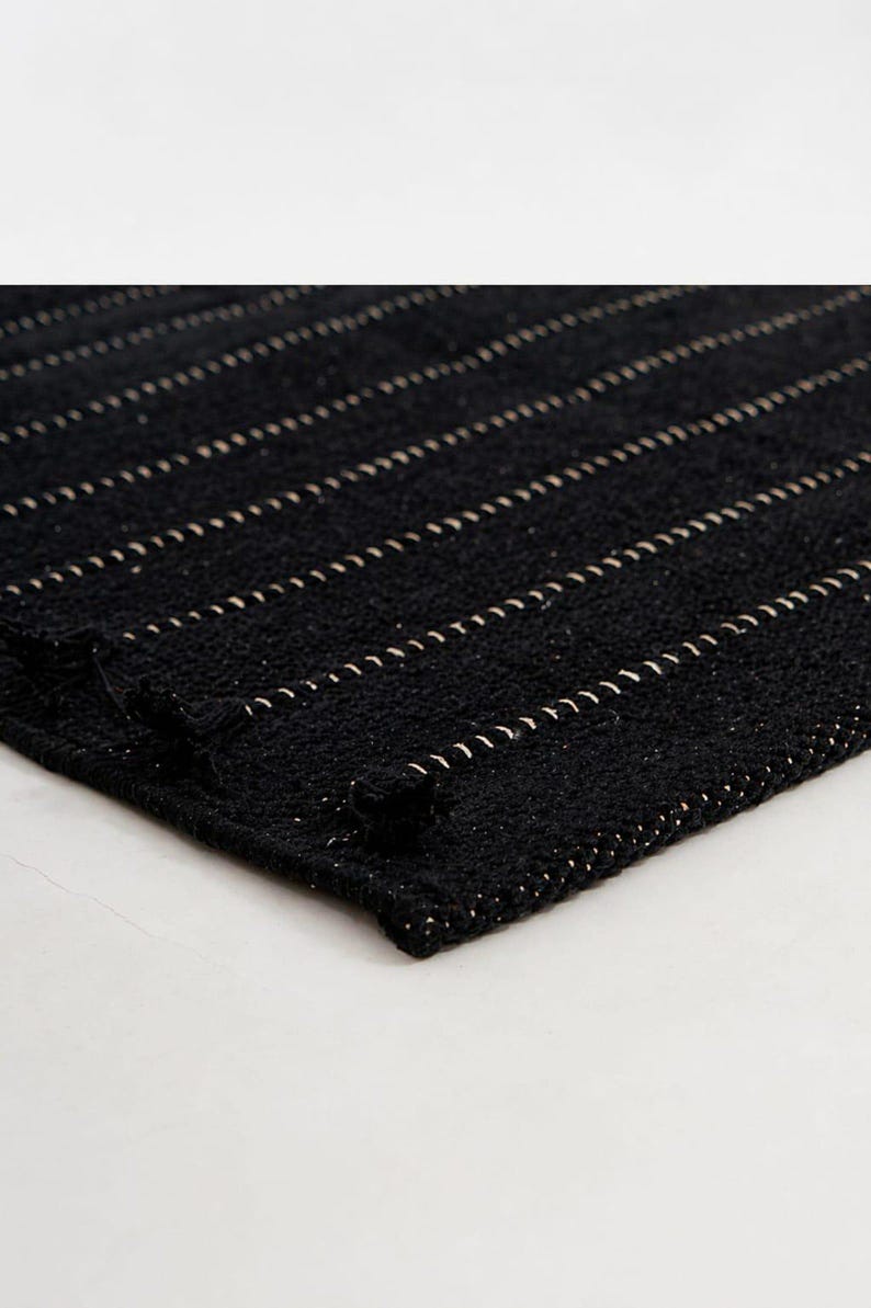 May include: A rectangular black woven rug featuring a series of parallel horizontal lines in a light beige or off-white color. The texture of the rug appears thick and coarse, with small knotted fringe details visible along the edge of the fabric.