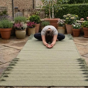 May include: A rectangular sage green woven mat featuring a textured surface with horizontal ribbed patterns and decorative fringe detailing along the short edges. The mat is laid out flat on a stone patio surface, showcasing its length and construction.
