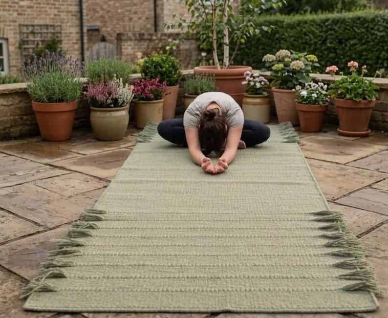 May include: A rectangular sage green woven mat featuring a textured surface with horizontal ribbed patterns and decorative fringe detailing along the short edges. The mat is laid out flat on a stone patio surface, showcasing its length and construction.
