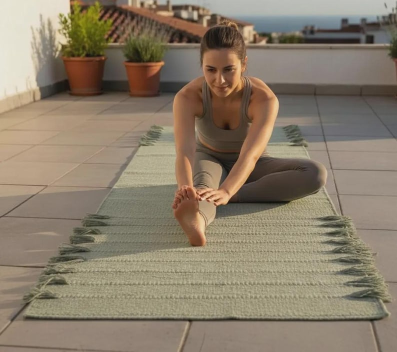 May include: A rectangular sage green yoga mat featuring a textured woven surface and decorative fringe detailing along both long edges. The mat is laid flat on a tiled surface, providing a stable base for stretching and various floor-based exercises.