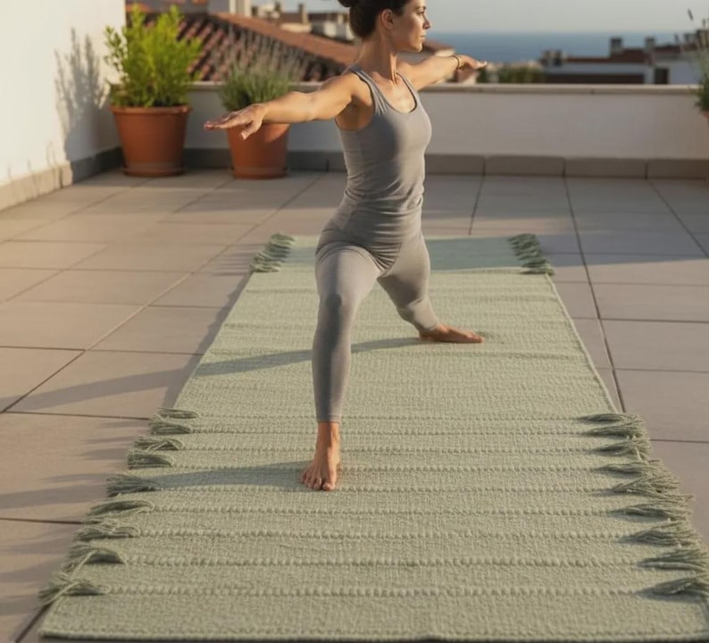 May include: A rectangular sage green yoga mat featuring a textured surface and decorative fringe tassels along both long edges. The mat is laid out flat, providing a stable base for exercise, with a consistent woven pattern throughout the material.