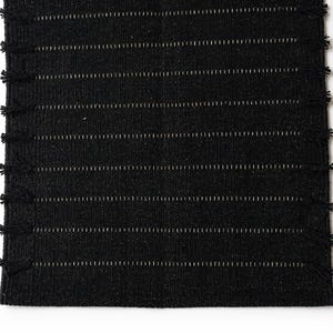 May include: A rectangular black woven table runner featuring a textured fabric surface with horizontal rows of thin, light-colored stitching. The edges of the runner are finished with small, evenly spaced black fringe tassels along both sides.