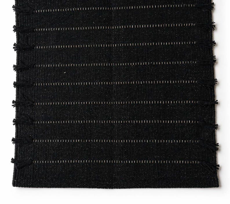 May include: A rectangular black woven table runner featuring a textured fabric surface with horizontal rows of thin, light-colored stitching. The edges of the runner are finished with small, evenly spaced black fringe tassels along both sides.