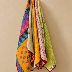 May include: A collection of colorful, patterned quilts hanging against a neutral wall. The quilts feature various designs, including floral, paisley, and geometric patterns, in shades of pink, yellow, blue, and green. Each quilt is trimmed with a contrasting red border.