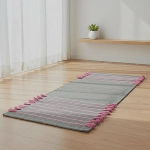 May include: A rectangular gray runner rug featuring thin horizontal pink stripes across the surface. The rug is finished with matching pink tassels attached along the two shorter ends, creating a textured border detail on the floor covering.