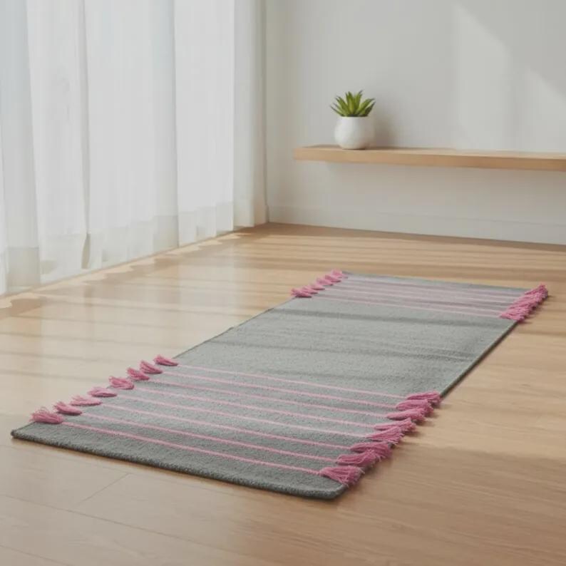 May include: A rectangular gray runner rug featuring thin horizontal pink stripes across the surface. The rug is finished with matching pink tassels attached along the two shorter ends, creating a textured border detail on the floor covering.