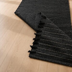 May include: A rectangular black woven rug with a folded corner revealing a textured underside. The rug features a series of small white horizontal dashes and a row of black fringe tassels along the edge of the folded section.