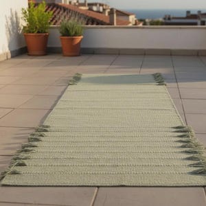 May include: A rectangular sage green runner rug featuring a textured woven surface and decorative fringe tassels along both of the shorter ends. The rug is laid out flat on a tiled surface, displaying a consistent light green color throughout.