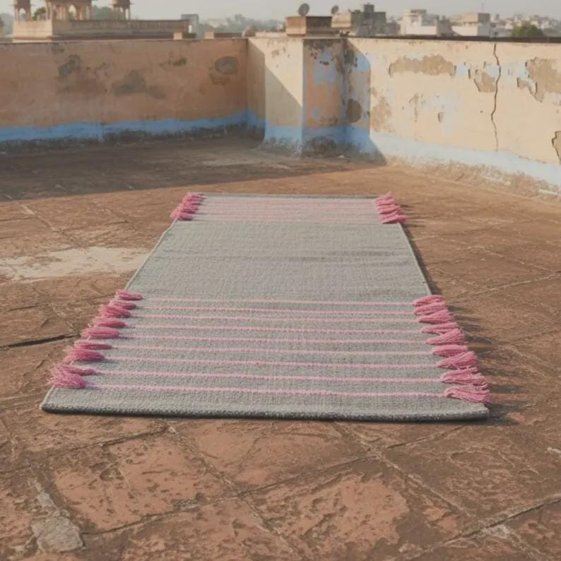 May include: A rectangular woven rug featuring a solid gray base with horizontal pink stripes spaced evenly across the surface. The rug is finished with decorative pink tassels attached along the two shorter ends of the textile piece.