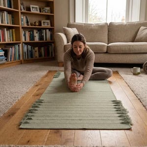 May include: A rectangular sage green woven mat featuring a textured surface with horizontal ribbed lines and decorative fringe detailing along the side edges, laid flat on a wooden floor surface for use as a floor covering or exercise accessory.