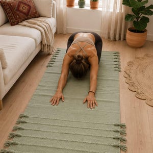 May include: A rectangular sage green yoga mat featuring a textured woven surface with horizontal ribbed detailing and decorative tassels along both short edges. The mat is crafted from a solid-colored fabric and provides a flat surface for exercise.