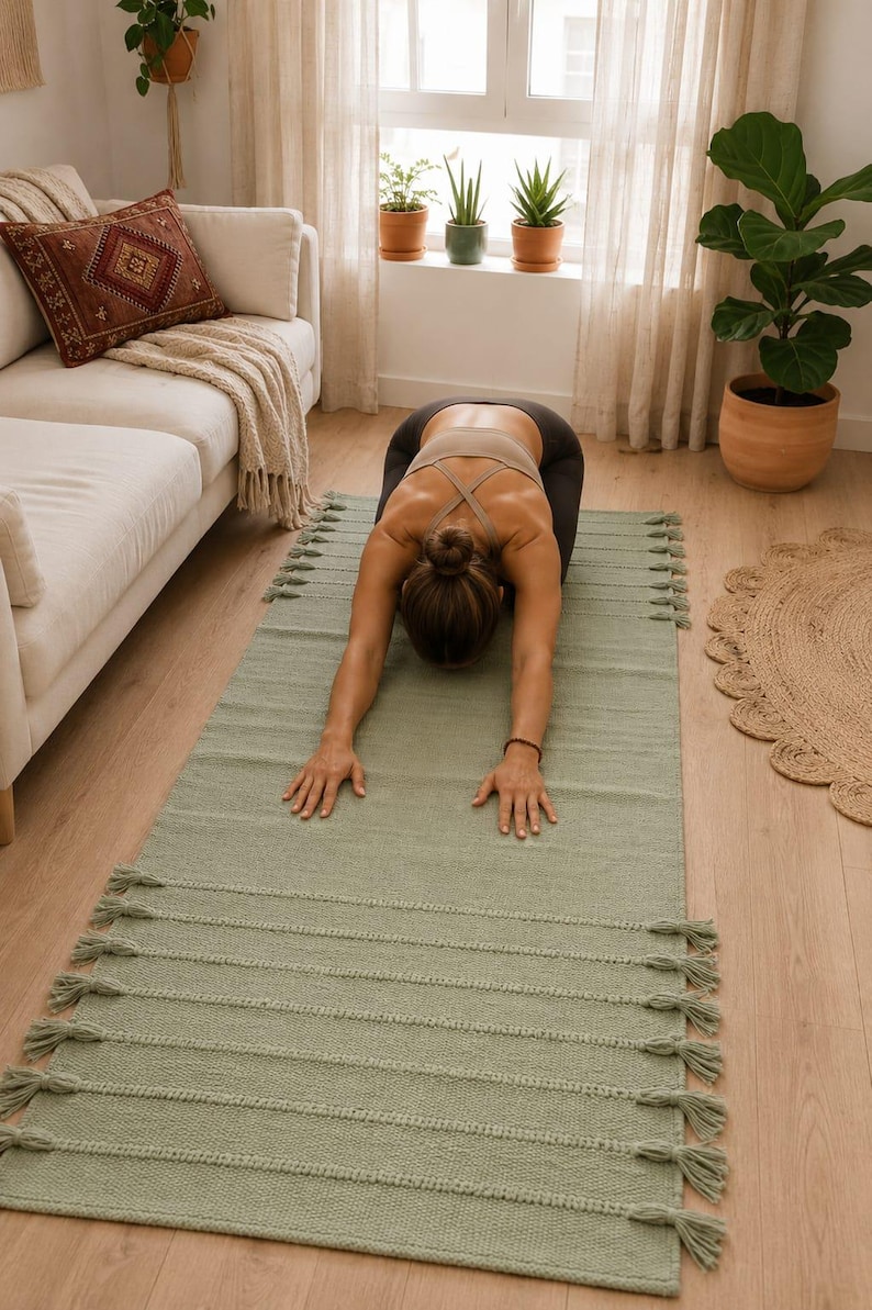May include: A rectangular sage green yoga mat featuring a textured woven surface with horizontal ribbed detailing and decorative tassels along both short edges. The mat is crafted from a solid-colored fabric and provides a flat surface for exercise.