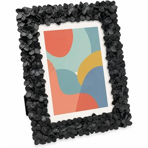 Könnte beinhalten: Schwarzer Bilderrahmen mit floralem Design. Der Rahmen umgibt einen farbenfrohen abstrakten Druck mit roten, blauen und gelben Formen. Der Rahmen besteht aus einem dunklen Material und hat einen eingebauten Ständer.
