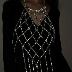 May include: A silver-toned metallic body chain featuring a diamond-shaped lattice pattern across the chest area, with long, dangling vertical strands extending downward from the base of the grid, worn over a plain black long-sleeved garment.
