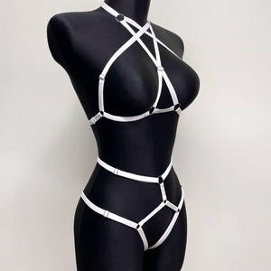 May include: A white strappy harness set featuring thin elastic bands, silver-toned metal rings, and adjustable sliders. The design includes a halter-style top piece connected to a waist and hip harness, creating a geometric pattern of straps.