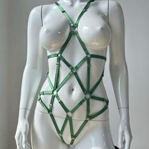 Puede incluir: Un arnés corporal verde construido con correas elásticas ajustables y anillos de metal plateado. El diseño presenta un patrón de red geométrico que cubre el torso y las caderas, con correas finas que se extienden sobre los hombros y la cintura.