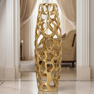 May include: A tall, gold-colored decorative vase with an open, organic, lattice-like design. The vase is cylindrical and stands on a reflective, tiled floor. The background includes a glimpse of a room with neutral-toned curtains and furniture.