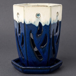 May include: A ceramic planter with a geometric design. The top section is white, transitioning to a deep blue with cut-out flame-like shapes. Includes a matching square base. The planter has a glossy finish and is suitable for indoor use.