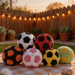 Puede incluir: Una colección de balones de fútbol de peluche de colores, incluyendo blanco y negro, rojo y negro, naranja y negro, rosa y blanco, amarillo y negro, y verde y amarillo. Las pelotas están dispuestas sobre una manta a cuadros al aire libre, con una valla de madera y luces de cadena en el fondo.