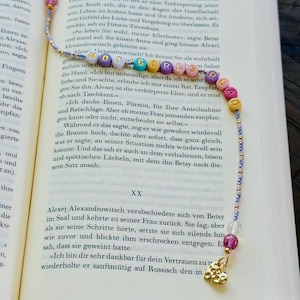 Könnte beinhalten: Ein Perlen-Lesezeichen mit dem Text "HOT GIRLS" in bunten Buchstabenperlen. Das Lesezeichen hat einen goldenen Anhänger und liegt auf einem offenen Buch.