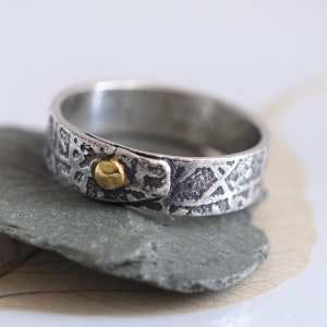 May include: A silver ring with a textured surface and a small gold accent. The ring is set on a grey stone, showcasing its unique design and craftsmanship. The band has a rustic, organic feel.