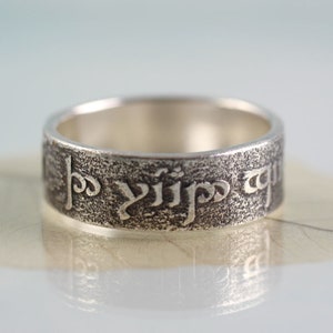 Könnte beinhalten: Ein silberner Ring mit einer Inschrift in einer alten Sprache. Die Inschrift lautet: "Ein Ring, sie alle zu beherrschen, Ein Ring, sie zu finden, Ein Ring, sie alle zu bringen und in der Dunkelheit zu binden."