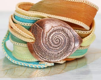Può includere: Un bracciale avvolgente con un elemento centrale in metallo color rame caratterizzato da un design in rilievo a forma di fossile di ammonite a spirale. Il cinturino è composto da nastri in tessuto multistrato nei toni arancio, ottanio e verde.