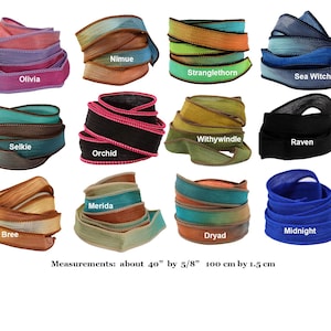 Op de afbeelding: Twaalf opgerolde stoffen linten in diverse kleuren en texturen, elk gelabeld met namen zoals Olivia, Nimue, Stranglethorn, Sea Witch, Selkie, Orchid, Withywindle, Raven, Bree, Merida, Dryad en Midnight. Elk lint meet 100 cm bij 1,5 cm.