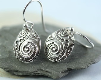 May include: A pair of silver-toned drop earrings. Each earring has a teardrop shape with an intricate design of spirals and dotted patterns. The earrings hang from a simple hook.