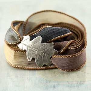 Könnte beinhalten: Ein gewickeltes Stoffarmband mit einem silberfarbenen Eichenblatt-Anhänger aus Metall. Das Band besteht aus geschichteten, strukturierten Stoffstreifen in Beige, Grau und Braun, die an den Rändern mit dekorativen Nähten versehen sind.