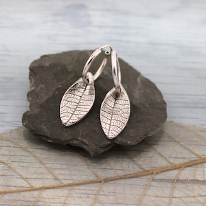 May include: Silver hoop earrings with leaf-shaped pendants. The pendants feature intricate vein detailing, resembling real leaves. The earrings are displayed on a dark gray stone against a light blue and gray background.