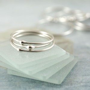 May include: Three silver rings with a rounded, teardrop-shaped detail on each ring. The rings are stacked on top of each other and are resting on a stack of frosted glass squares.