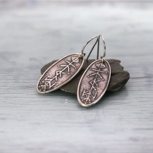 May include: A pair of silver earrings with an oval shape and a geometric design. The earrings are hanging from a silver hook.