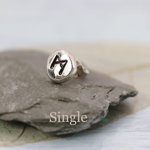 May include: A single silver stud earring featuring a black rune symbol. The earring has a textured surface and is shown on a grey stone. The word "Single" is printed in white below the earring.
